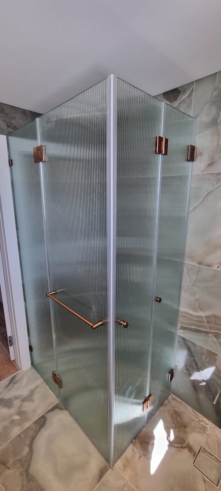 מקלחון פינתי מזכוכית ריבס עם צירים בגוון נחושת על רקע שיש ירקרק
